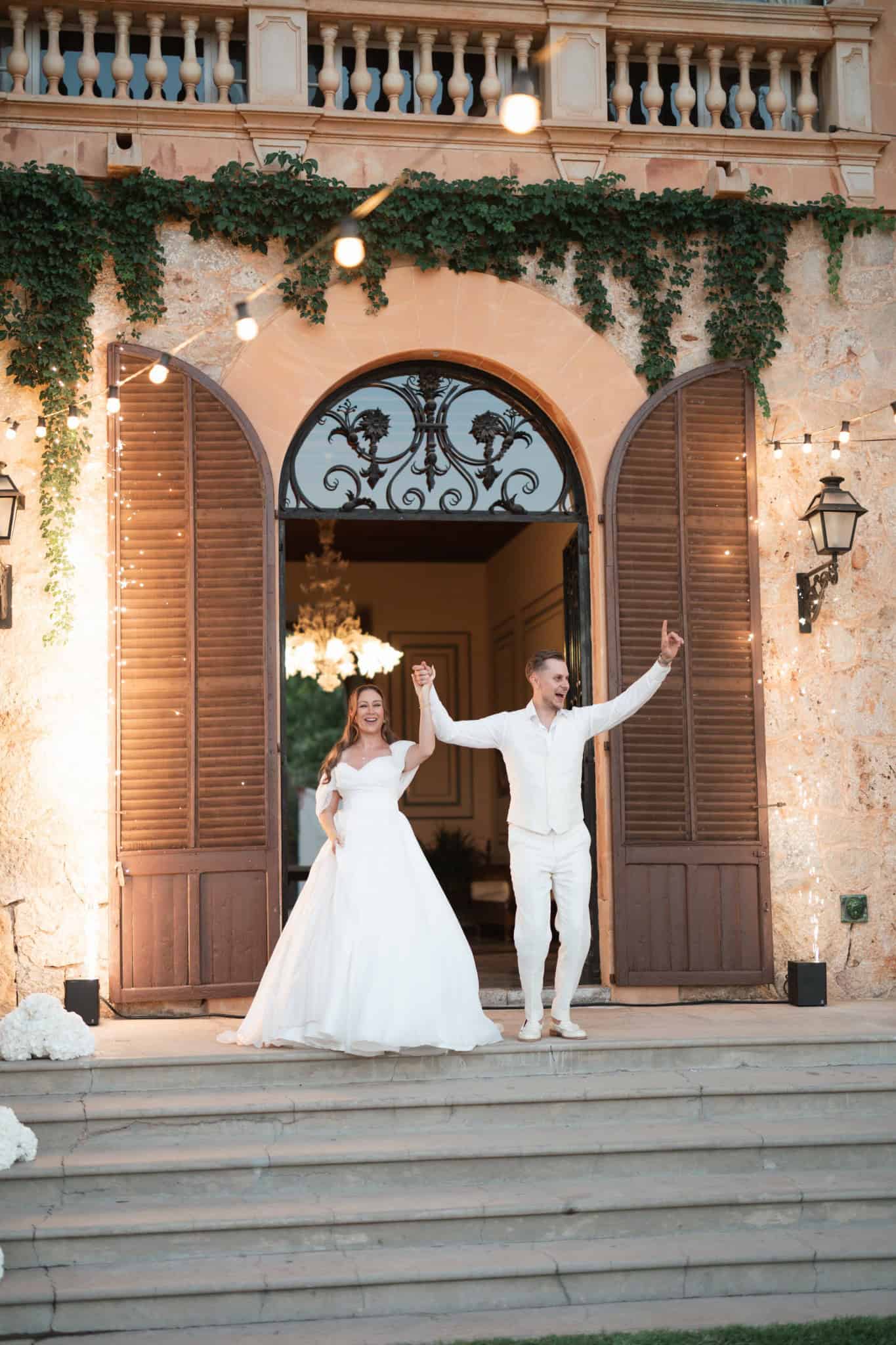 Yaiza and Stefan beside an arched stone façade with wooden shutters and climbing greenery at Son Togores.