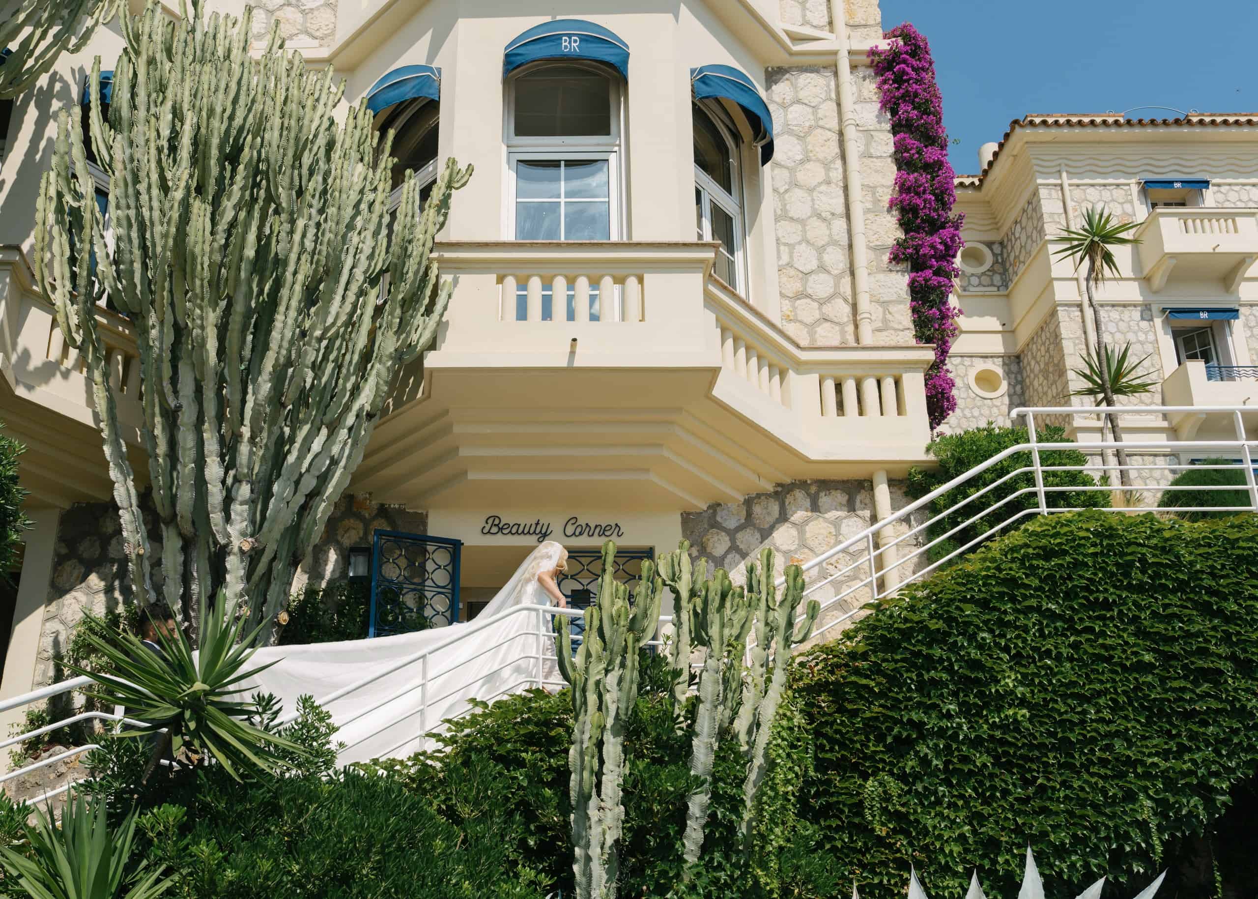 Bride on the steps of Hôtel Belles Rives beneath blue awnings and flowering bougainvillea.