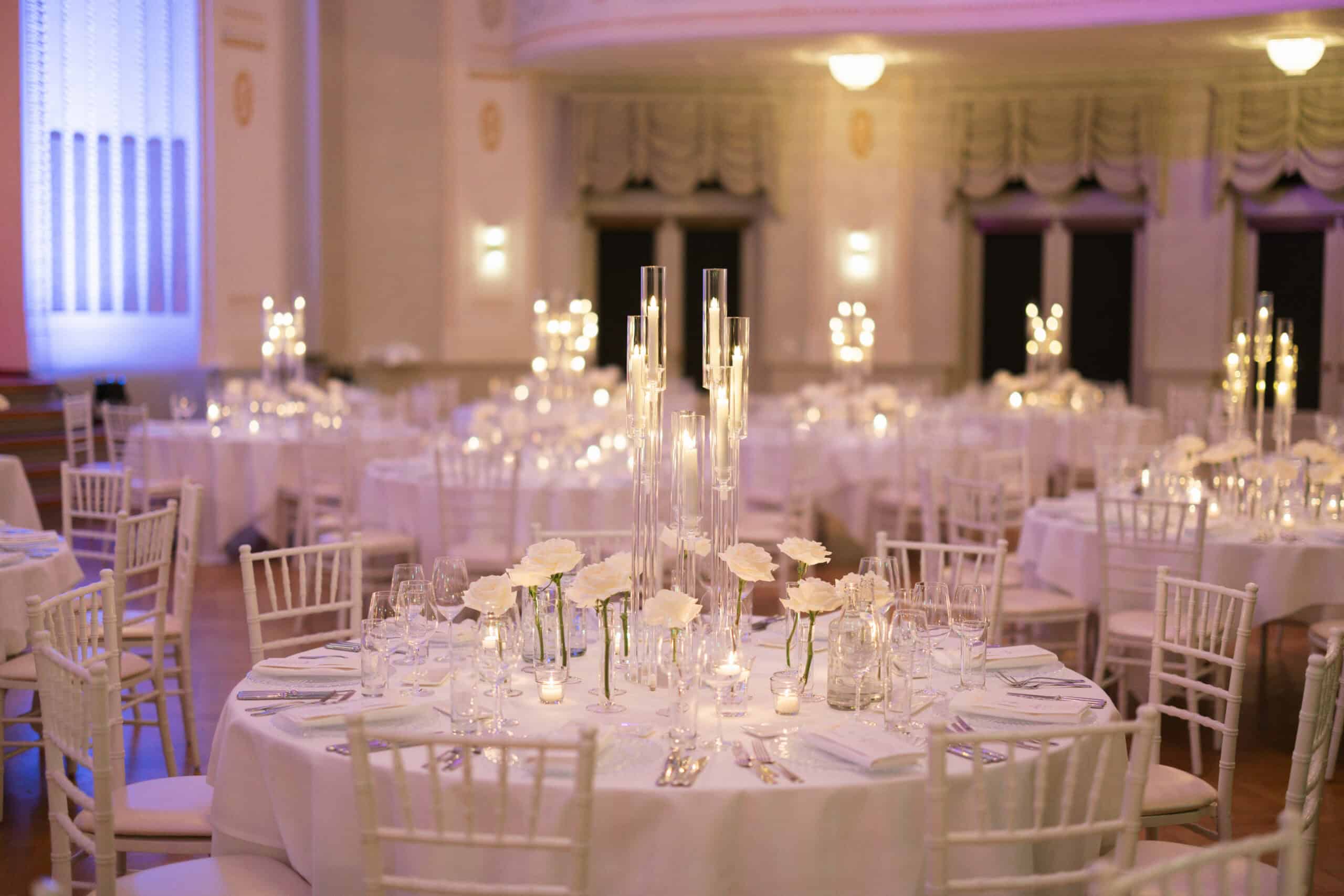 Reception table with white linen, white flowers in glass vases and candles in tall glass cylinders.