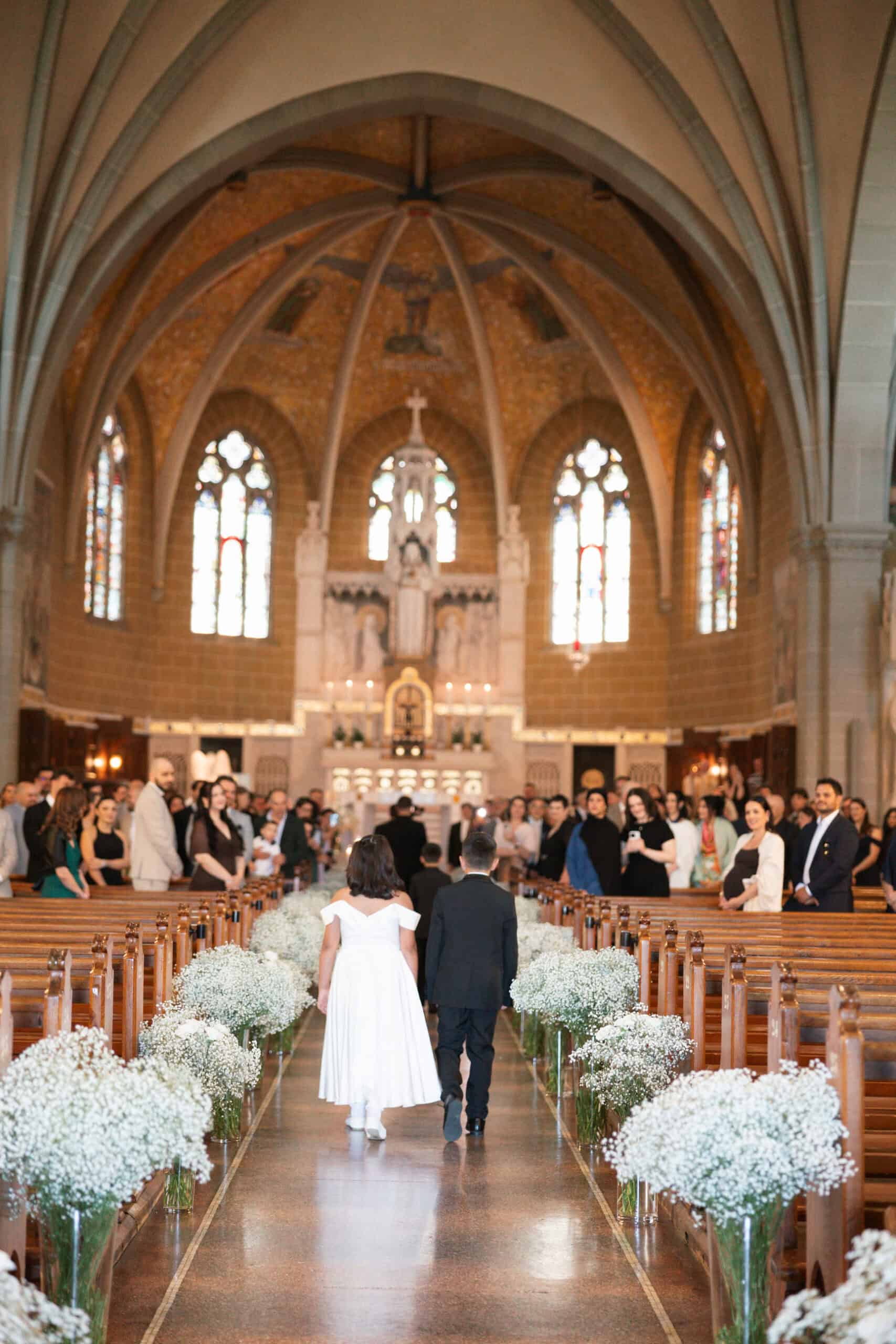 Francesca and Giuseppe walking through a church aisle in Zug, lined with white flowers beneath tall arches.