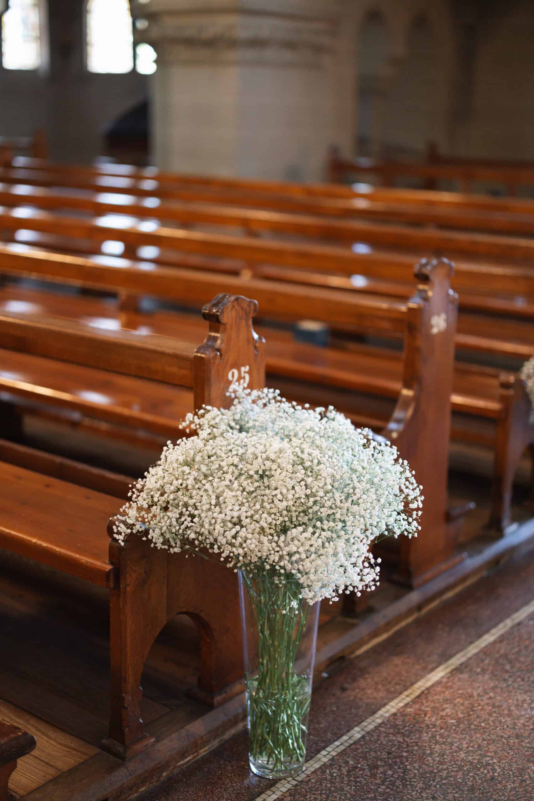White flowers and baby’s breath in a glass vase beside a wooden church pew.