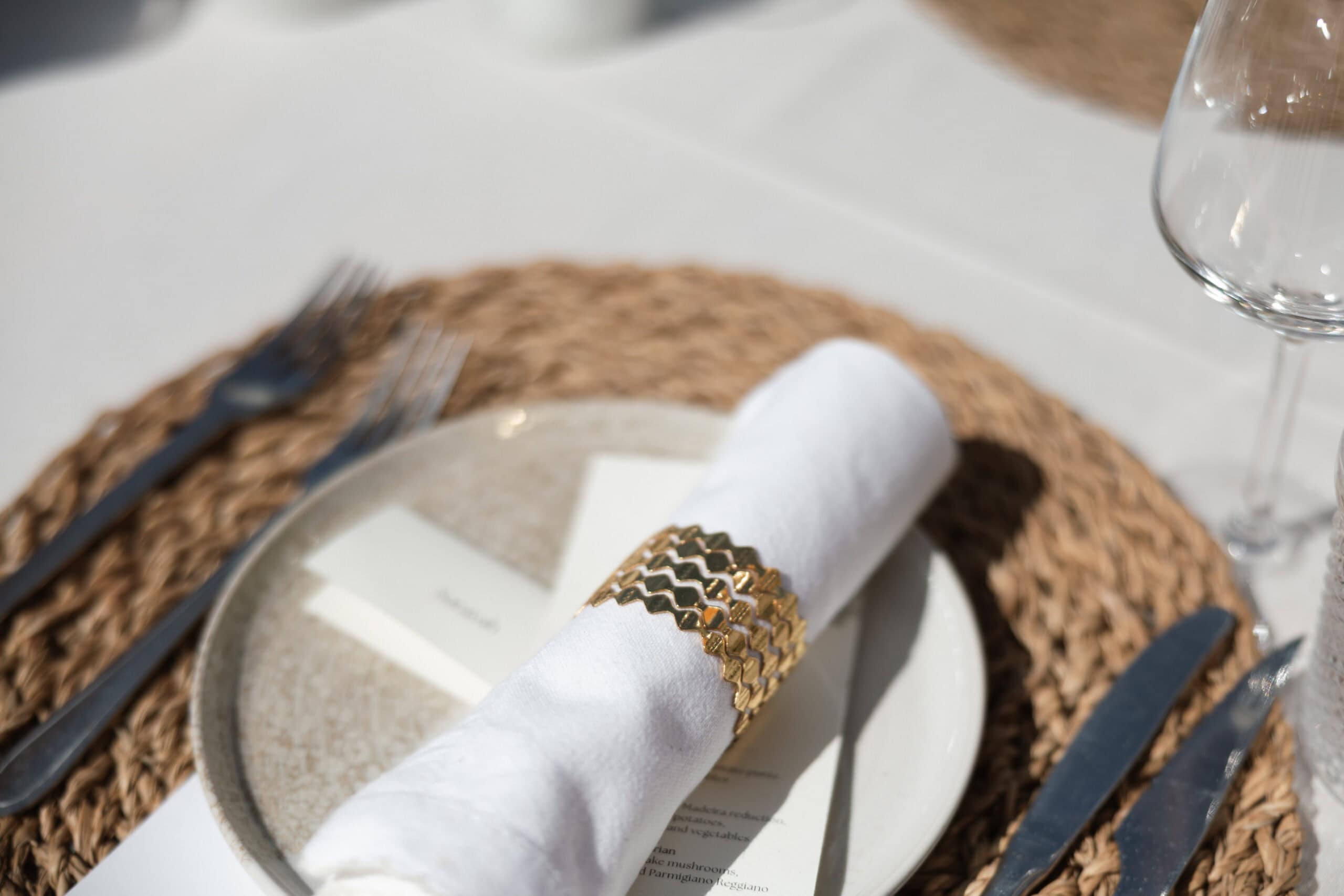 Wedding place setting with a woven placemat, rolled white napkin and sculptural gold-toned napkin ring.