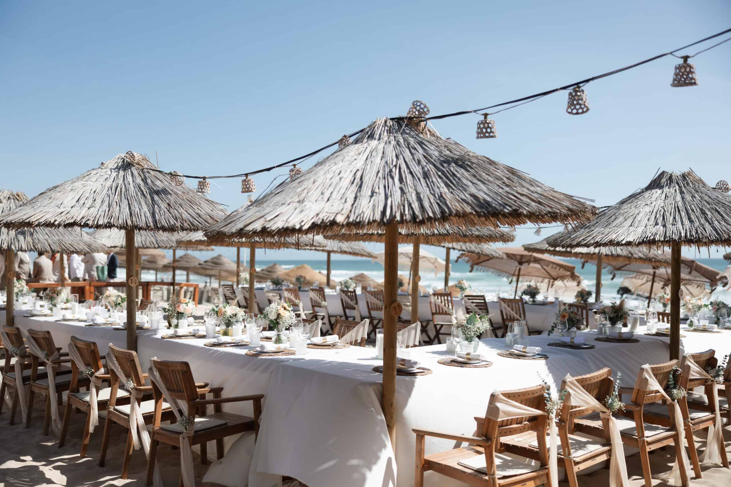 Coastal wedding reception in Costa da Caparica, Portugal, with long tables, thatched parasols and an ocean view.